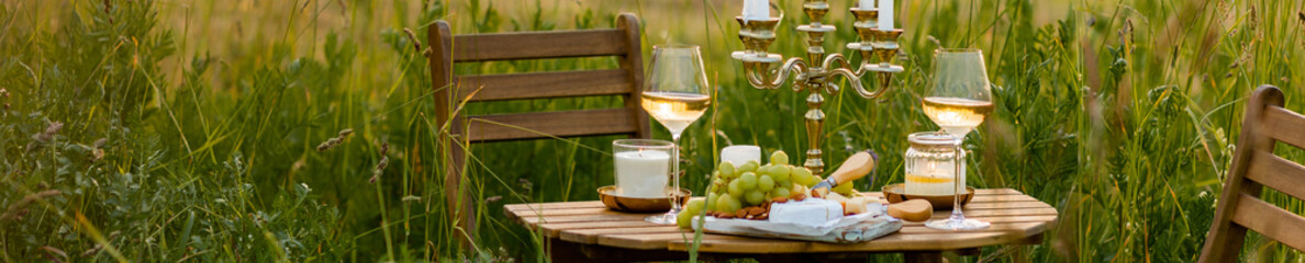Surprise romantic date outdoors in a meadow. Elegant table with snacks and wine, chandelier with candles as decor. Private party celebration of special event for loving couple. Banner