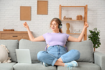 Young woman in headphones with laptop meditating on sofa at home