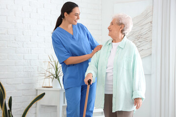 Physical therapist helping senior woman with stick to walk in bedroom