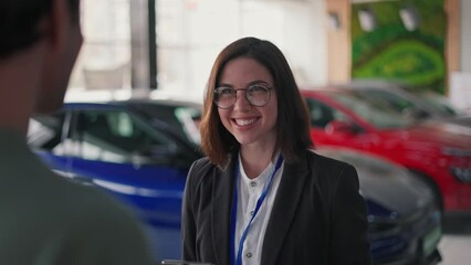 Over the shoulder, a confident brunette girl with glasses in a business suit communicates with a guy client in a car showroom. Girl manager communicates with a client in a car showroom