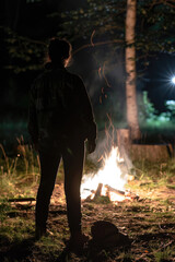 Fototapeta premium A person stands near a crackling campfire in the woods at night. Their silhouette is visible against the bright flames, creating a stark contrast