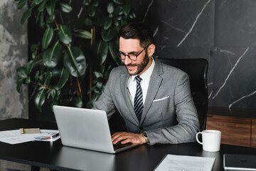 Successful boss typing on laptop remotely in office. Young rich manager businessman checking financial data, doing managerial online job, receiving official letters and e-mails