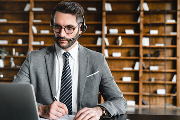 Successful young salesman wearing headphones consulting customers online on laptop. Businessman e-learning, watching webinar and taking notes, studying foreign language, watching tutorial