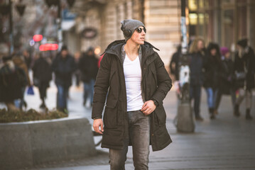 Portrait of Handsome Smiling Young Man with Woolen Hat, Sunglasses and Earphones on the Street Listening to Music