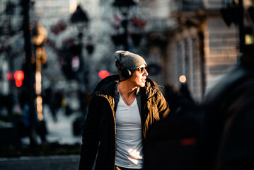 Portrait of Handsome Smiling Young Man with Woolen Hat, Sunglasses and Earphones on the Street...