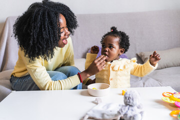 African mother feeding grapes to toddler while playing with toys on the couch