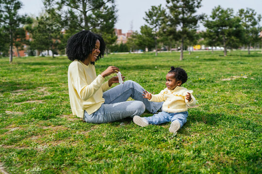 A woman and a child are sitting on the grass in a park with blowing bubbles