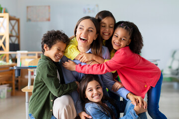 Happy diverse kids, junior school children embracing female teacher appreciating and thanking educator in classroom