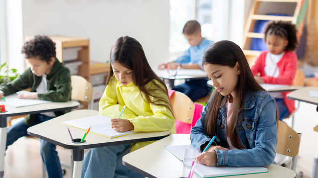 Education concept. Group of diverse preteen students writing school test, noting lesson in their copybooks, sitting at desks in classroom - Powered by Adobe
