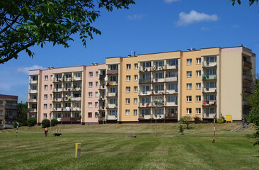 Four-story low building. A restored 1980s block of flats in a sunlit estate, next to a grassy field...