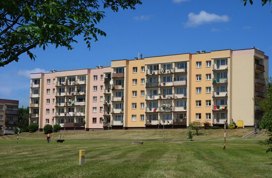 Four-story low building. A restored 1980s block of flats in a sunlit estate, next to a grassy field with recently mown grass. 