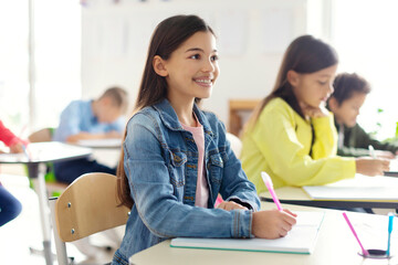 Smiling cute European schoolgirl in casual attentively listening to teacher while sitting at her...