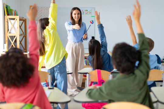 Students raising their hands up in classroom at international elementary school. Cheerful female teacher playing with pupils