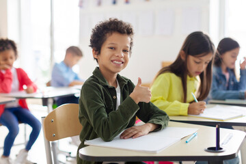 Portrait of smiling Latin schoolboy showing thumbs up and smiling, sitting at desk in his classroom with classmates on background.