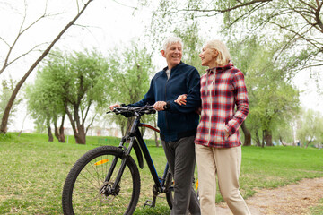 elderly senior couple rides bicycle in the park in the summer and smiles, old gray-haired man and woman are actively resting outdoors, old people practice cycling in forest