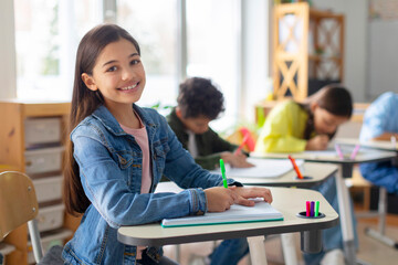 Happy smart schoolgirl sitting at school desk in classroom on background with classmates friends, looking and smiling at camera