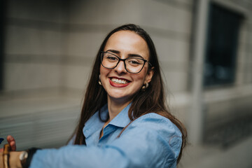 Confident businesswoman smiling brightly, wearing glasses and a casual blue shirt, in an outdoor...