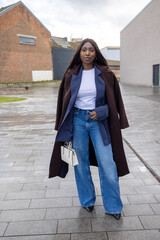 Fototapeta premium Chic street style: Woman in a brown coat, white top, blue jeans, holding a handbag on a rainy urban day.