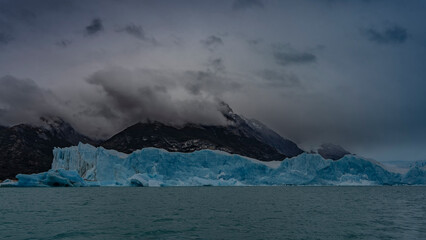 A wall of blue ice across a turquoise glacial lake. Icebergs, melted ice floes float in the water. Mountains in clouds and fog. Perito Moreno glacier. El Calafate. Argentina. Lago Argentino. 