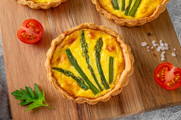 Mini tarts with green asparagus and cheese with cream filling on a wooden board on a light concrete background. Top view, selective focus.