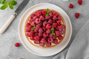 Delicious raspberry tart with vanilla custard cream sprinkled with powdered sugar on a white plate on a gray concrete background. Summer dessert.