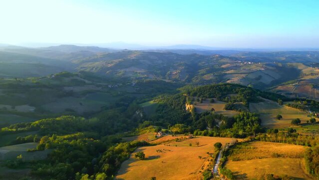 Steep aerial push-in and pan-right shot from Penna San Giovanni (MC) flying over brown fields, sunlit green thickets in the golden hour, roads stretching along and extensive hills in the far distance