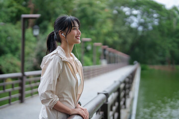 Young asian sporty women standing near lake with happiness and wearing earphones to listening music for resting after jogging outdoor in the park and fitness exercise for healthy wellness lifestyle