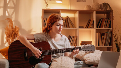 Online guitar class. Virtual music. Talented musician woman learning playing string instrument on laptop at home living room interior with free space.