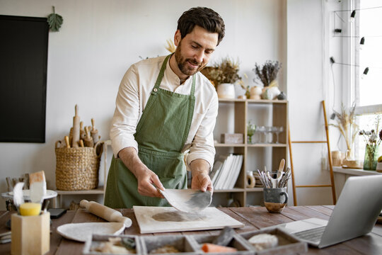 Artist is shaping clay and interacting through laptop, surrounded by tools and pottery materials. Potter wearing apron creating ceramic vase while participating in an online workshop at home studio.