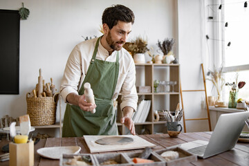 Artist is shaping clay and interacting through laptop, surrounded by tools and pottery materials. Potter wearing apron creating ceramic vase while participating in an online workshop at home studio.