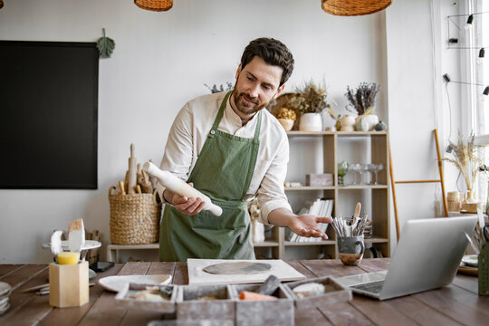 Artist is shaping clay and interacting through laptop, surrounded by tools and pottery materials. Potter wearing apron creating ceramic vase while participating in an online workshop at home studio.