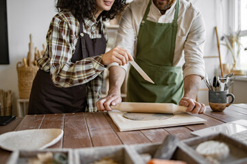 Obraz premium A man rolls clay with a rolling pin while a woman stands next to him holding a stack. They are working together in a home pottery studio, showcasing teamwork, creativity, and craftsmanship.