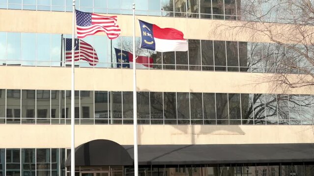 American and North Carolina state flags waving in the wind outside a modern office building with reflective windows.