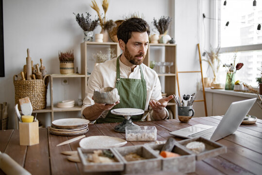 Bearded male studying remotely at university sitting in pottery studio. Handsome mature male potter in apron holding online lesson on clay vase sculpting using laptop.