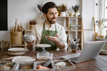 Bearded male studying remotely at university sitting in pottery studio. Handsome mature male potter in apron holding online lesson on clay vase sculpting using laptop.