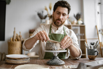 Smiling man working with clay in cozy home pottery studio. Bearded male seated at wooden table with various tools, materials, and laptop in bright well-decorated room.