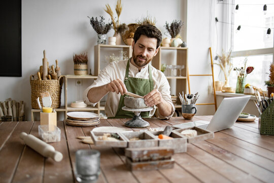 Bearded male seated at wooden table with various tools, materials, and laptop in bright well-decorated room. Smiling man working with clay in cozy home pottery studio.
