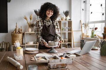 Smiling woman working on pottery in her studio, surrounded by tools and laptop. Creative and...