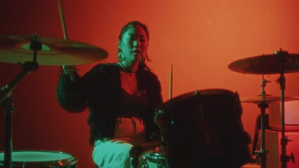 Young beautiful Asian girl in trendy outfit playing drum kit during solo music performance in studio with bright red background and neon illumination
