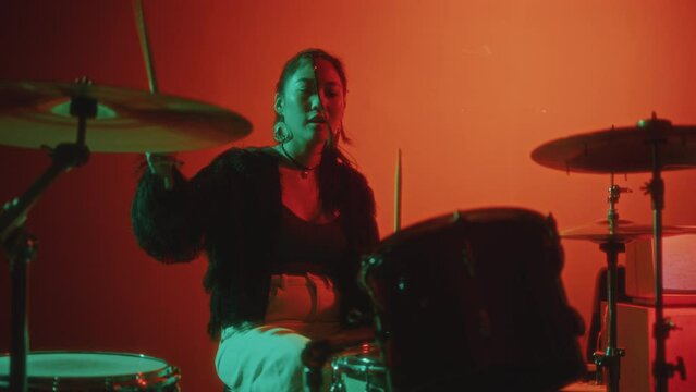 Young beautiful Asian girl in trendy outfit playing drum kit during solo music performance in studio with bright red background and neon illumination