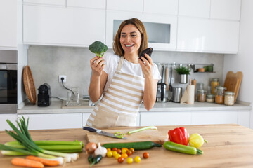 Photo of young woman smiling with red bell pepper while cooking and making salad with fresh...