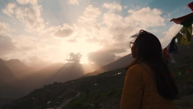 Silhouette of teenager Indian girl looking at dramatic sunset sky in mountains at Lahaul, Himachal Pradesh, India. Orange sky with sunrays bursting from clouds. Travel and holidays concept. 