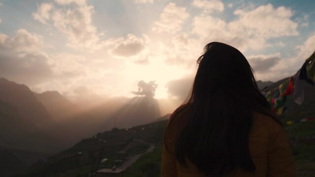 Silhouette of young Indian woman hopefully looking at sunset sky in mountains at Lahaul, Himachal Pradesh, India. Dramatic sunset sky with orange clouds and sunrays. Hope and faith concept.