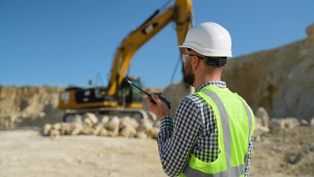Foreman in uniform giving commands to employee in excavator using walkie talkie at construction site