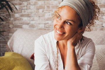Happy curly woman sitting on sofa at home smiling happily. Portrait of comfortable middle-aged lady in spring clothes relaxing in living room. Portrait of beautiful female smiling and dreaming