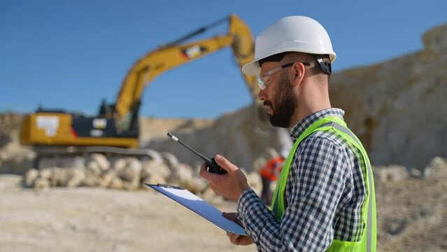 Engineer in uniform giving commands to employee in excavator using walkie talkie at construction site