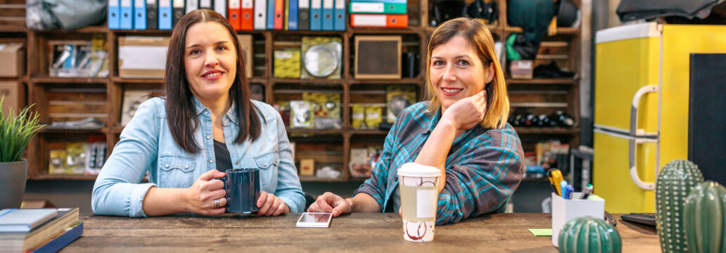 Portrait of two smiling female entrepreneurs looking at camera behind of counter in local motorbike shop. Banner of happy women colleagues working in store selling spare parts. Small business concept