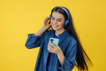 Smiling happy young woman wearing casual denim shirt dancing happily while listening music in wireless headphones over yellow background.