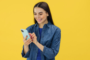 Adorable caucasian woman in casual clothes standing with a smartphone in her hand while buying online or typing message isolated over yellow background.