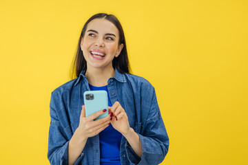 Young adult smiling happy pretty woman holding mobile phone device using apps on cellphone standing isolated on yellow background.